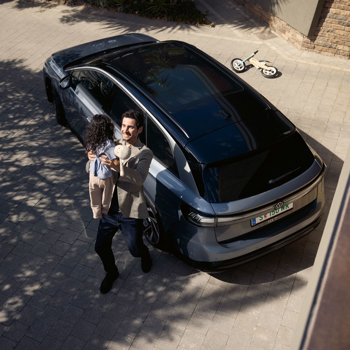 Mann hebt sein Kind hoch, was eine Puppe in der Hand halt und dahinter steht geparkt in der Einfahrt ein ID.7 Tourer und ein umgekipptes Kinderfahrrad.