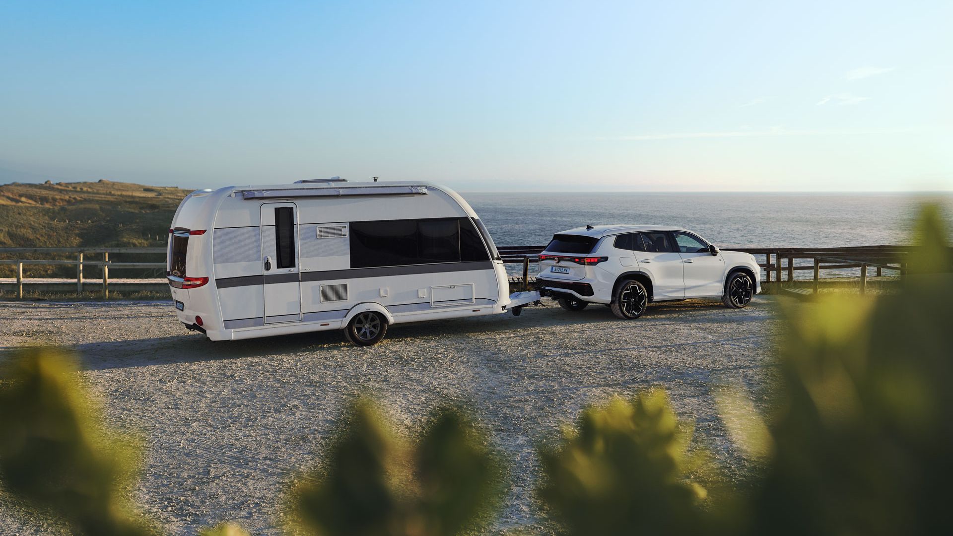 Weißer VW Tayron mit angehängtem Wohnwagen parkt auf einer Schotterfläche mit Meerblick und Dünen im Hintergrund.