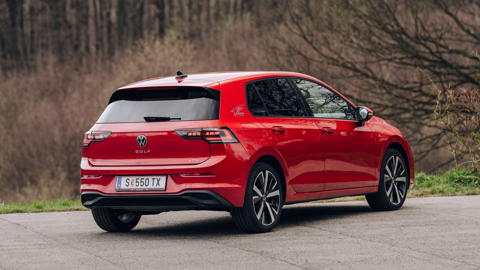 Roter VW Golf Kompaktwagen, leicht schräg von hinten fotografiert, geparkt auf einer asphaltierten Fläche. Im Hintergrund sind kahle Bäume und eine leicht verschwommene, natürliche Umgebung zu sehen. Das Auto wirkt modern und sportlich, mit dunklen Fenstern und markanten Rückleuchten.