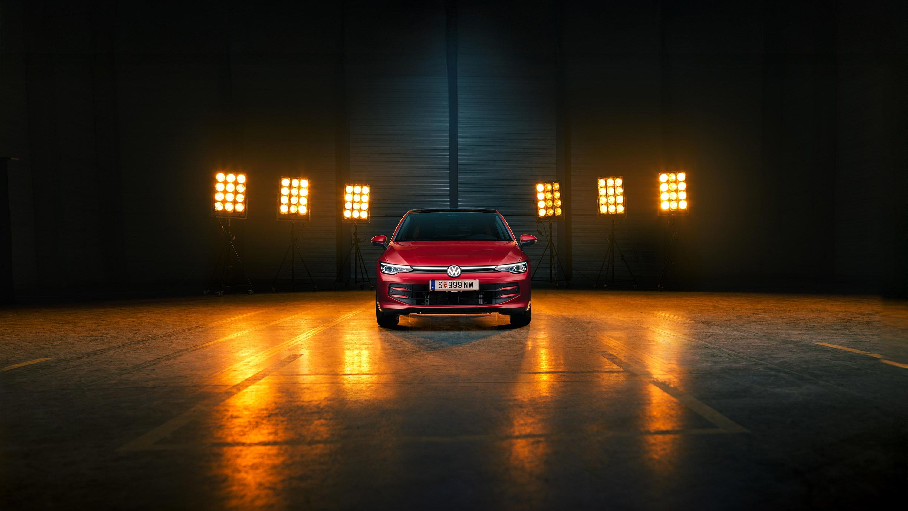 Ein roter VW Golf Falco Edition in Frontalansicht steht mittig im Bild in einer leeren Halle, Bühnenlichter strahlen das Fahrzeug von hinten an.