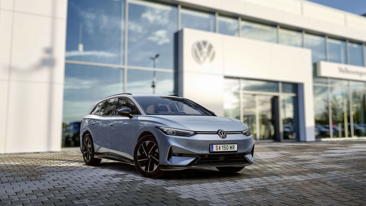 Ein silberner Volkswagen ID.7 Tourer steht auf einem gepflasterten Parkplatz vor einem modernen Volkswagen-Gebäude. Das Gebäude hat große Fenster und reflektiert den blauen Himmel. Rechts im Bild ist etwas Grün zu sehen.