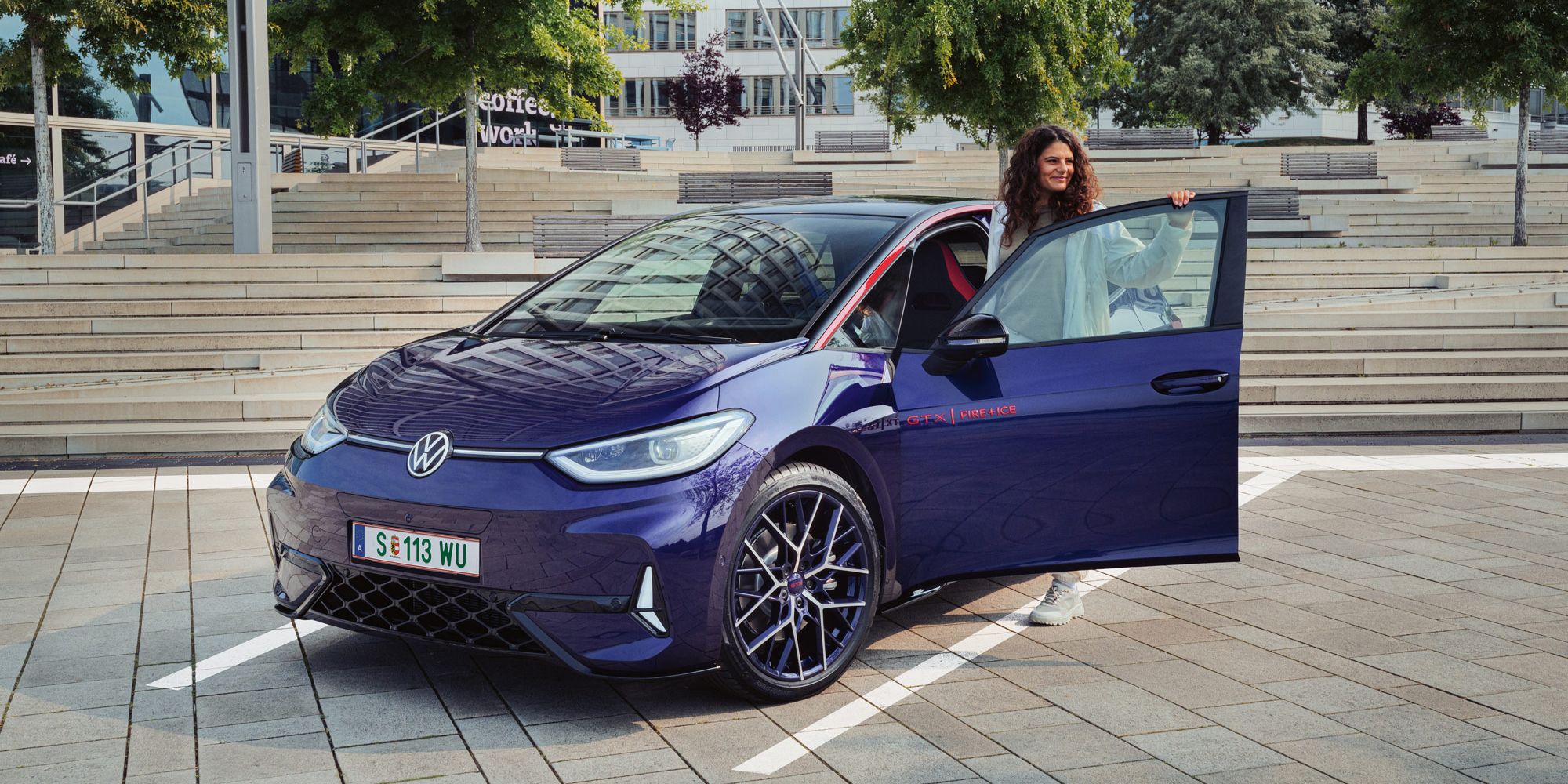 Eine Person steigt aus einem dunkelblauen VW Elektroauto auf einem modernen Parkplatz aus. Im Hintergrund sind Treppen und grüne Bäume zu sehen. Im Vordergrund des Autos sind Markierungen auf dem Asphalt sichtbar.