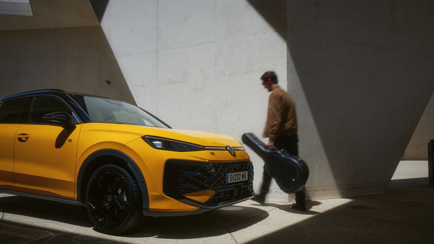 Ein Mann mit Gitarrenkoffer geht an einem gelben VW T-Roc R-Line in moderner Architektur vorbei.