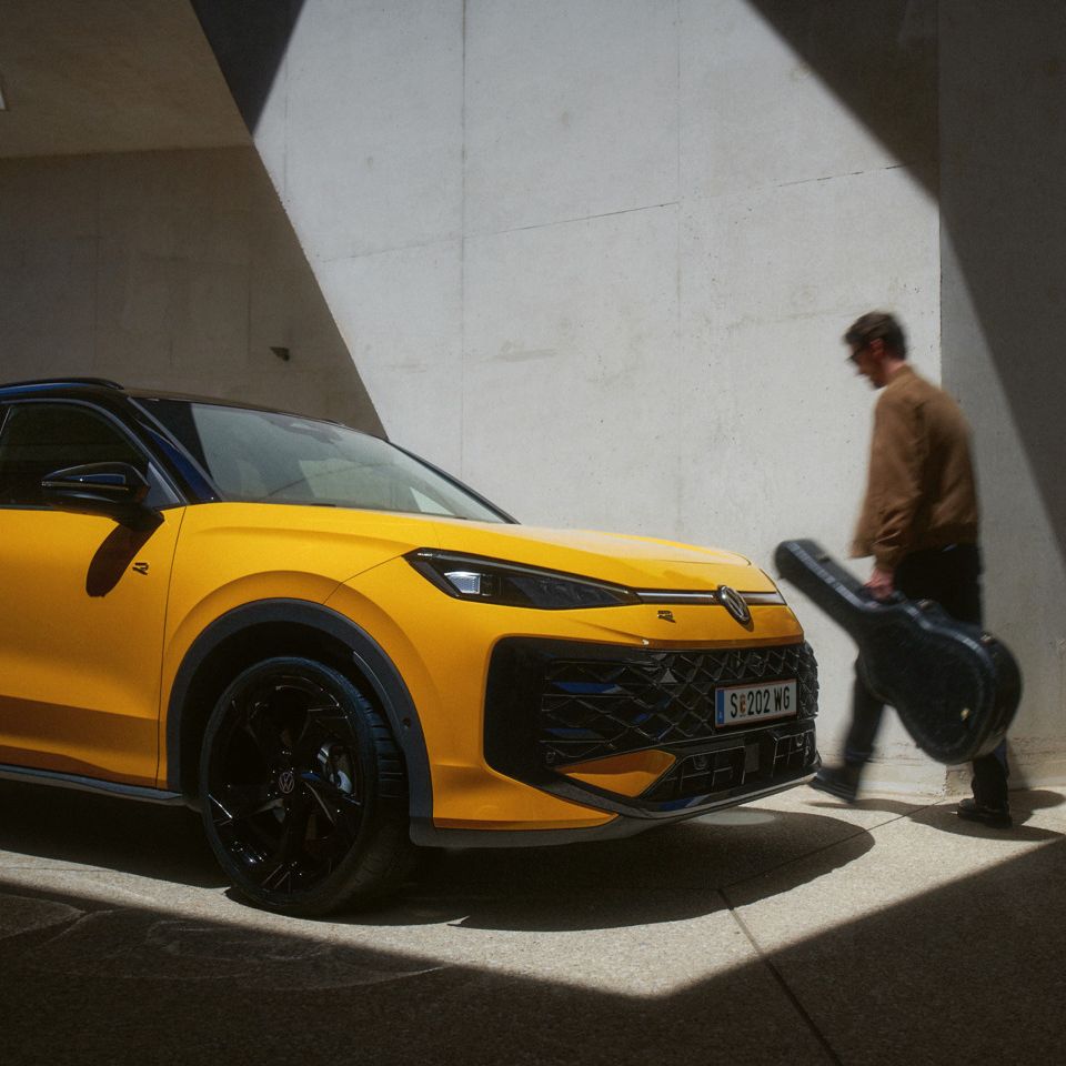 Ein Mann mit Gitarrenkoffer geht an einem gelben VW T-Roc R-Line in moderner Architektur vorbei.