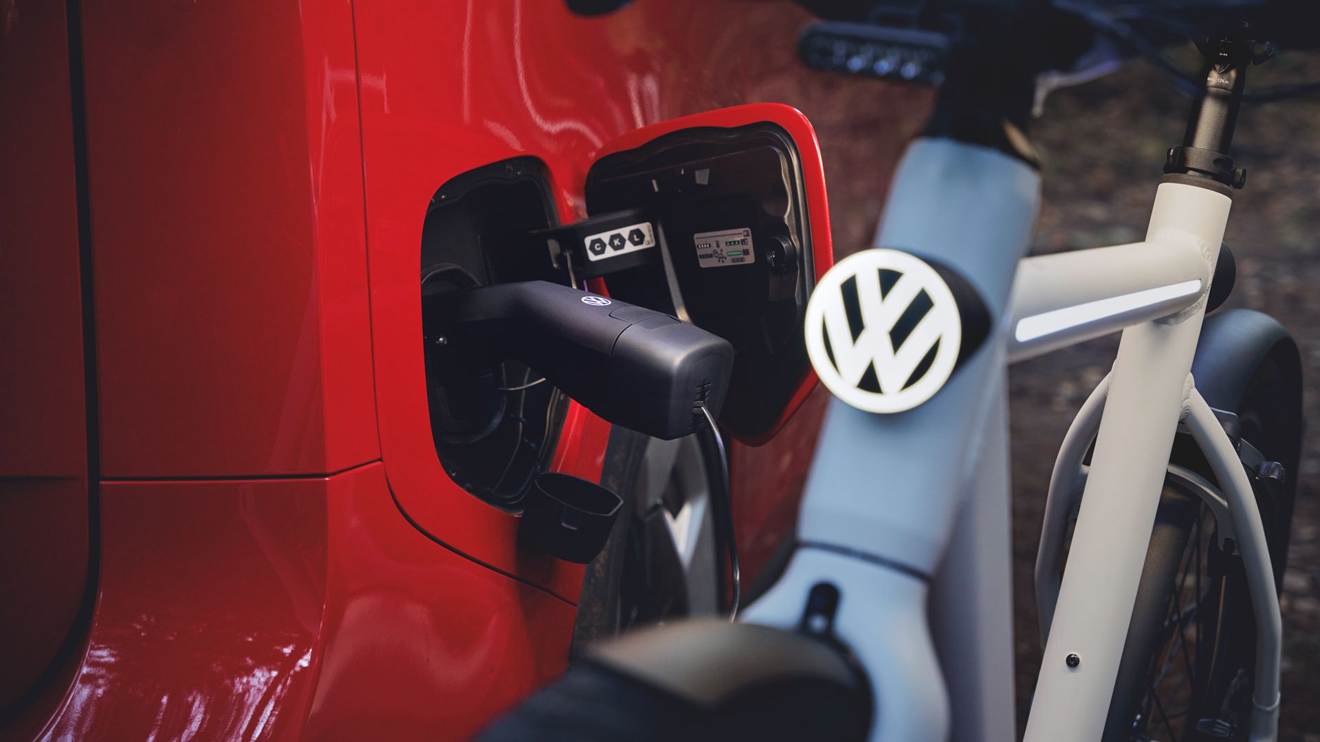 Nahaufnahme eines roten Elektroautos, das über ein Ladekabel mit einer Ladesteckdose verbunden ist. Im Vordergrund steht ein weißes E‑Bike mit einem gut sichtbaren VW-Logo auf dem Lenker. Die Szene vermittelt nachhaltige Mobilität mit Auto und Fahrrad.