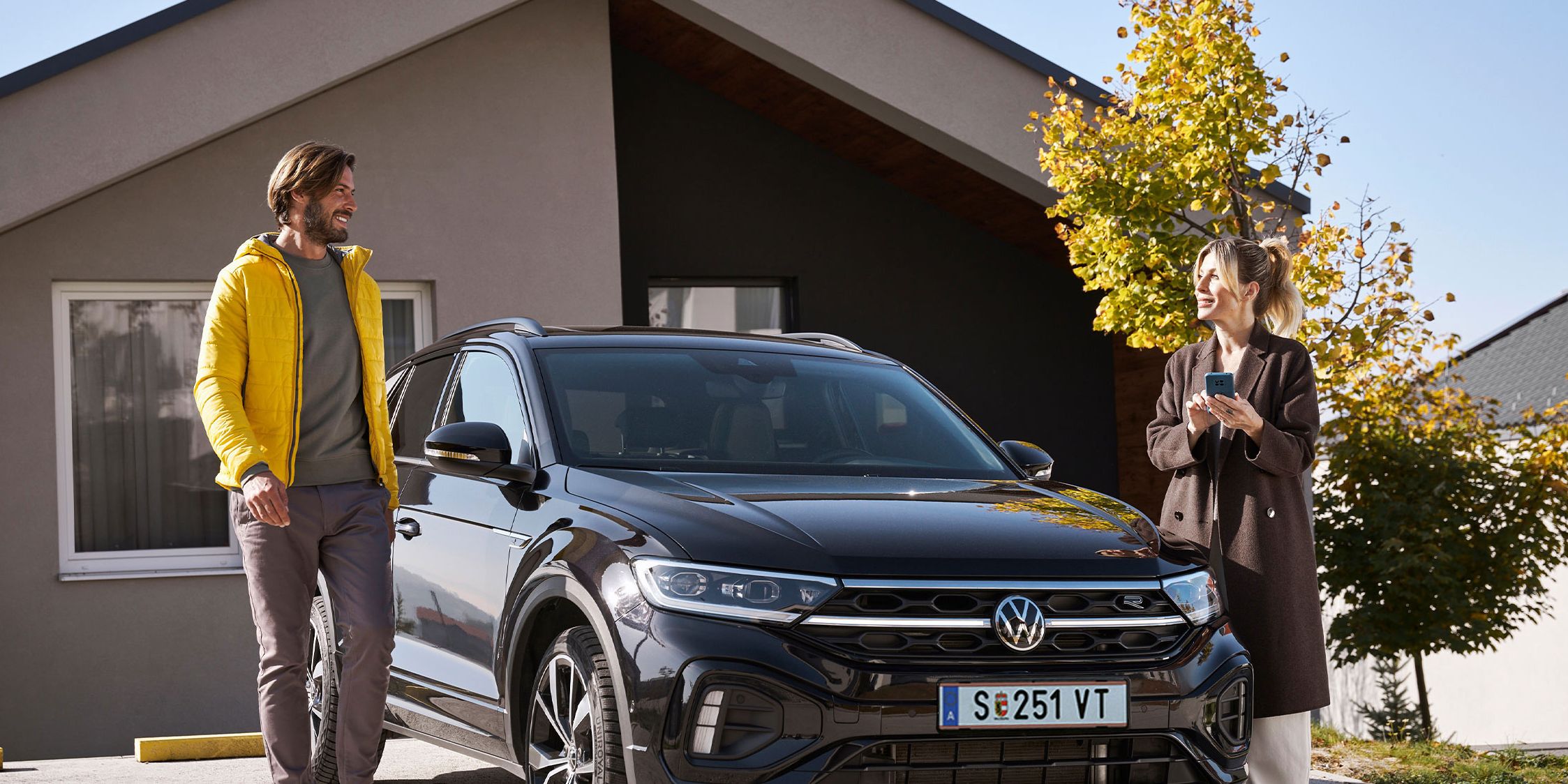 Mann und Frau steht mit Smartphone neben einem geparkten T-Roc vor einem Haus.