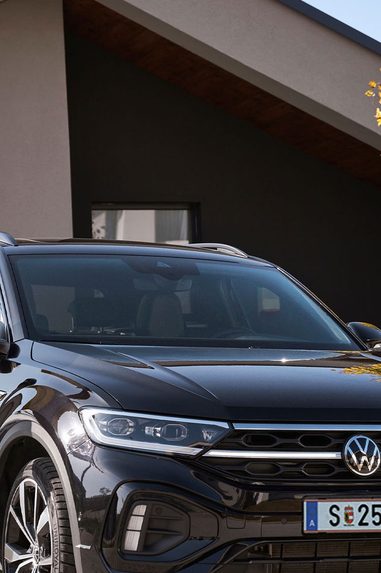 Mann und Frau steht mit Smartphone neben einem geparkten T-Roc vor einem Haus.