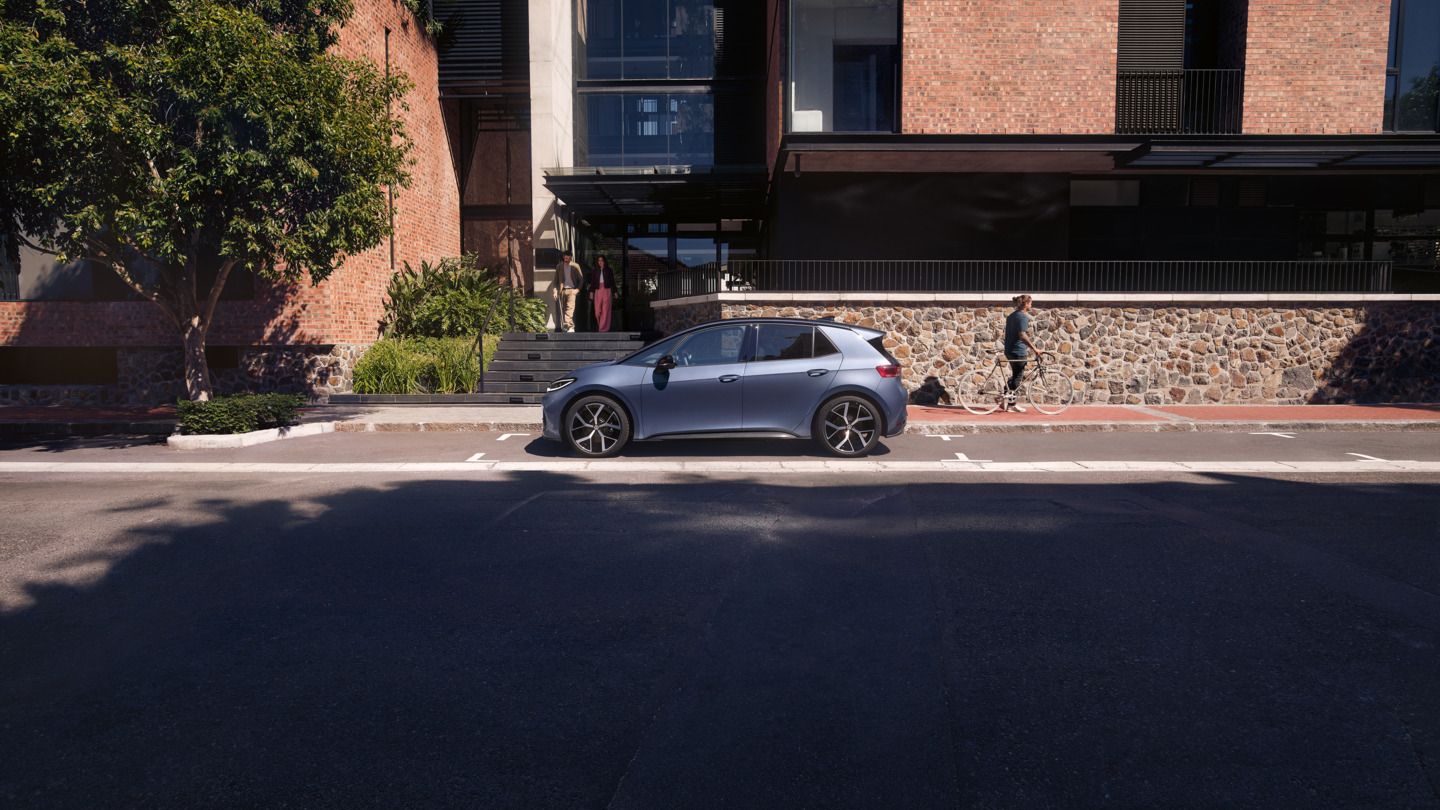 Ein blauer VW ID.3 Neo parkt vor einem Wohngebäude, aus dem eine Frau und ein Mann kommen