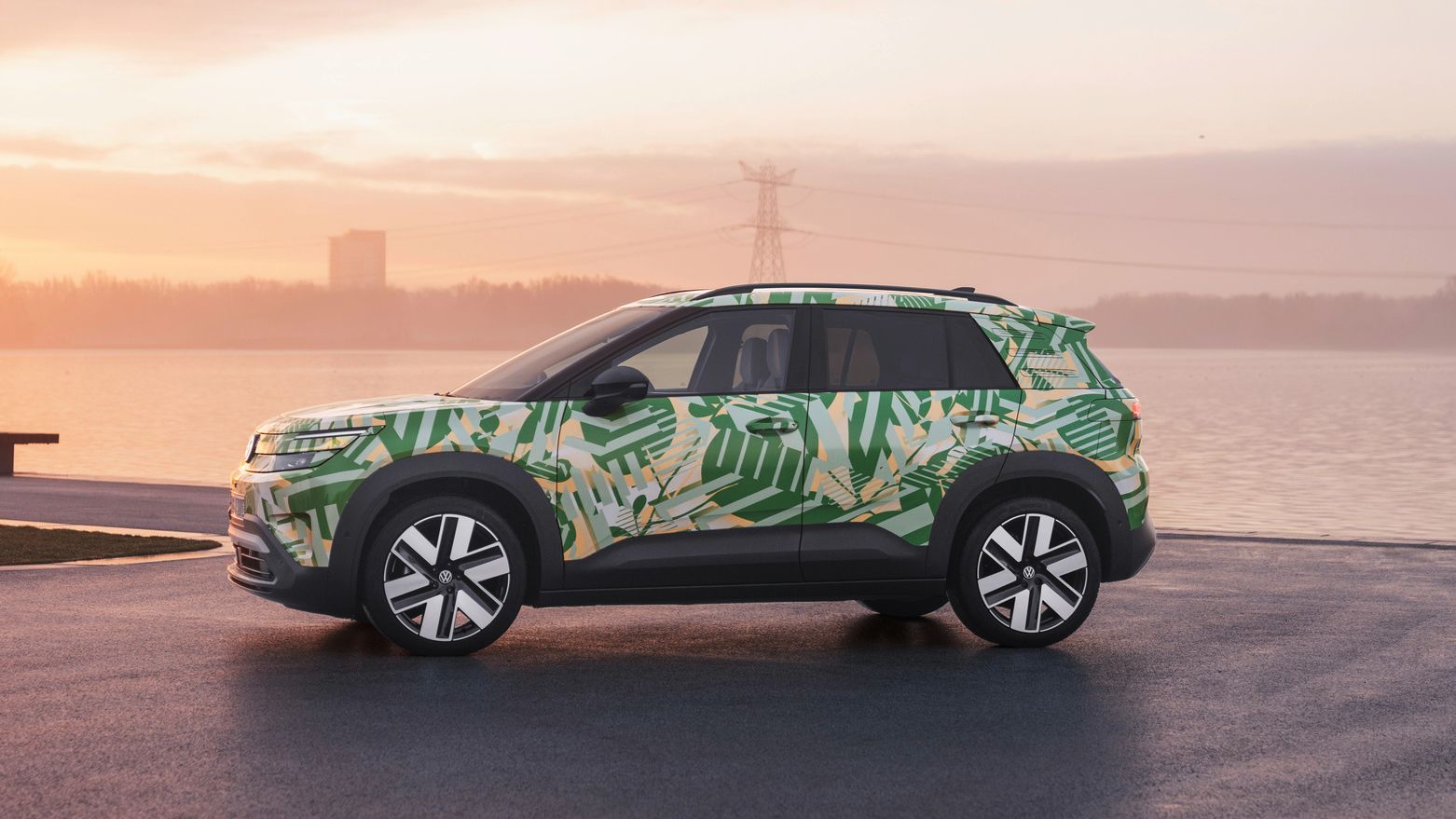 Ein SUV mit einer grünen und weißen Tarnlackierung steht auf einem asphaltierten Platz vor einem Gewässer bei Sonnenuntergang. Im Hintergrund sind Strommasten und Gebäude zu sehen. Der Himmel ist leicht bewölkt und in warmen Farben gehalten.