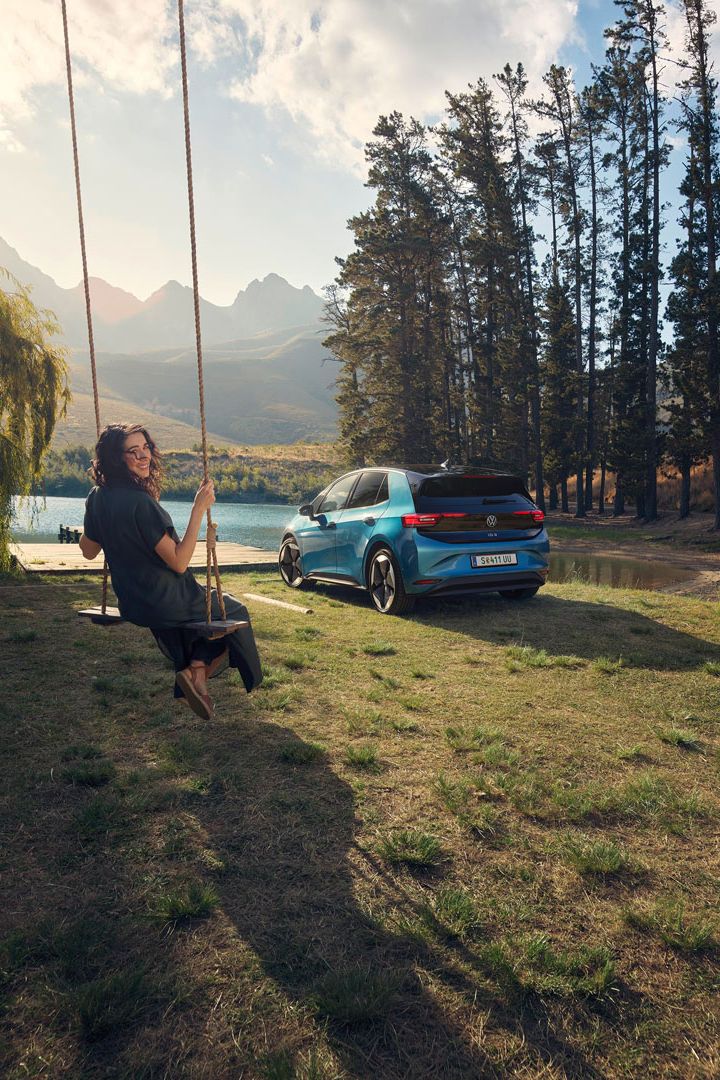 Person schaukelt auf einer Schaukel in einer landschaftlich reizvollen Gegend mit Bergen und einem See im Hintergrund. Neben der Person steht ein blaues Auto vor einer Reihe von Bäumen. Die Szene vermittelt ein Gefühl von Freizeit und Natur.
