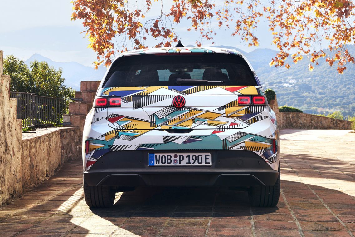 Ein ID. Polo steht auf einer gepflasterten Terrasse mit Blick auf eine bergige Landschaft. Das Fahrzeug hat ein farbenfrohes, geometrisches Muster und ein deutsches Kennzeichen. Im Hintergrund sind herbstliche Bäume und ein antikes Gebäude zu sehen.