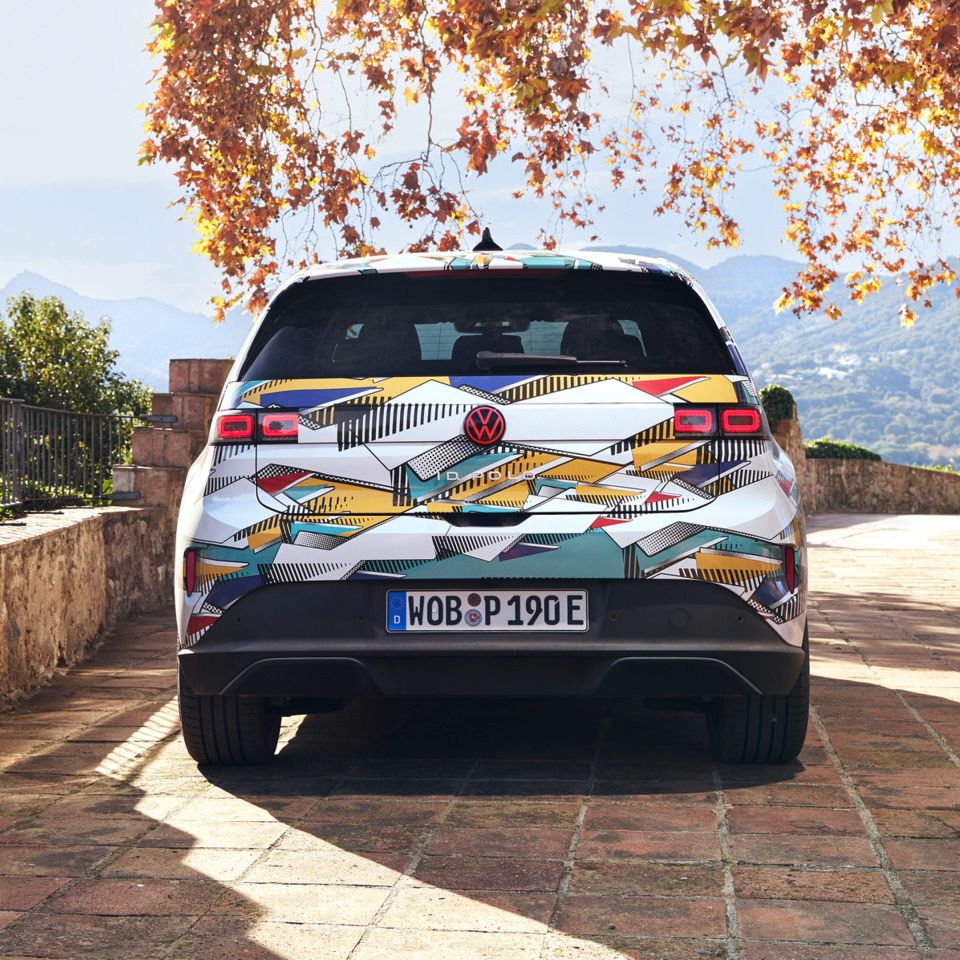 Ein ID. Polo steht auf einer gepflasterten Terrasse mit Blick auf eine bergige Landschaft. Das Fahrzeug hat ein farbenfrohes, geometrisches Muster und ein deutsches Kennzeichen. Im Hintergrund sind herbstliche Bäume und ein antikes Gebäude zu sehen.