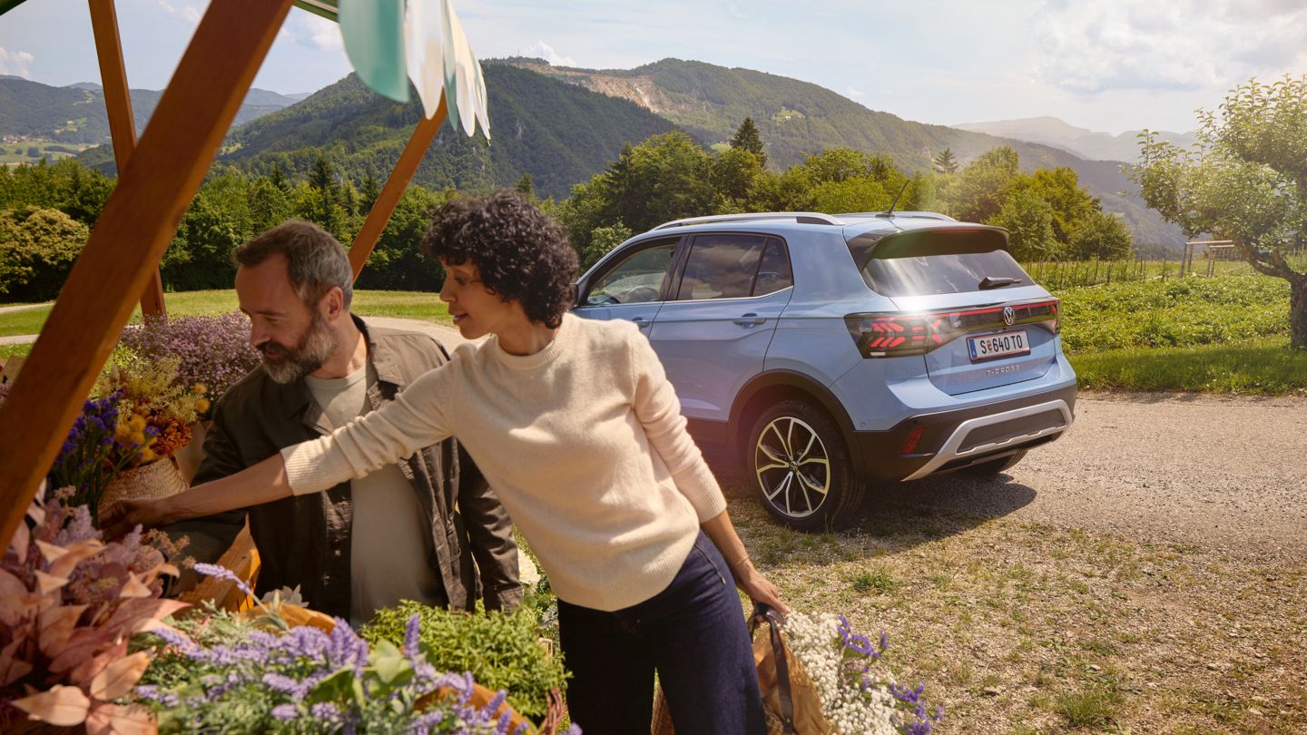 Mann und Frau stehen bei einem Blumenstand, hinter ihnen ein blauer VW T-Cross vor bergiger Landschaft.