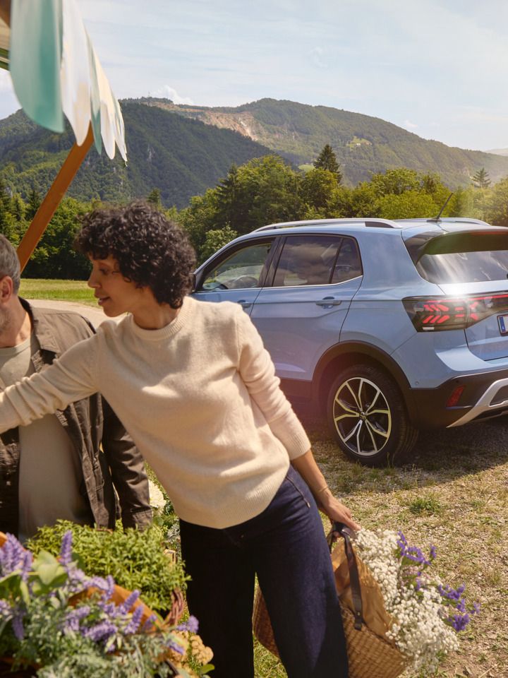 Mann und Frau stehen bei einem Blumenstand, hinter ihnen ein blauer VW T-Cross vor bergiger Landschaft.