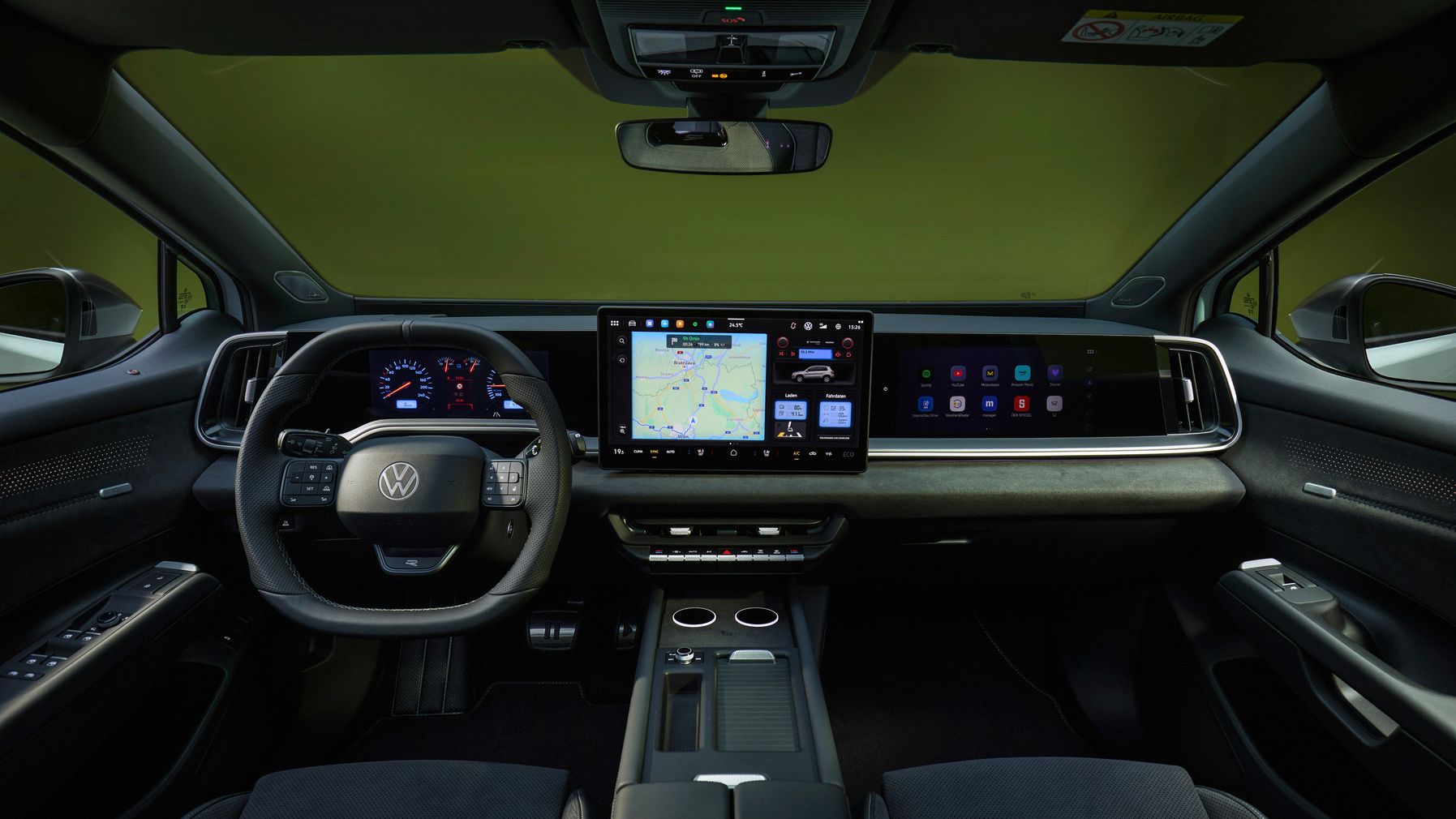 Innenraum eines modernen Volkswagen mit Fokus auf Cockpit und Armaturenbrett, aufgenommen von den Rücksitzen aus. Vorne befindet sich ein digitales Kombiinstrument hinter dem Multifunktionslenkrad und ein großer zentraler Touchscreen mit Navigationskarte, daneben ein weiterer breiter Bildschirm für Infotainment. Die Mittelkonsole bietet Getränkehalter und Ablagefächer, das Interieur wirkt aufgeräumt und hochwertig.