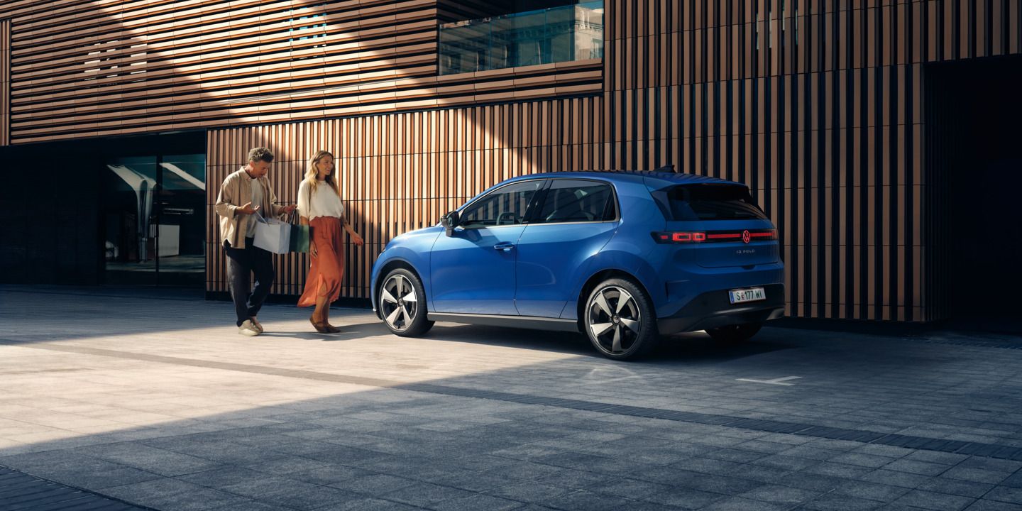 Ein blauer VW ID. Polo Style steht vor einem Gebäude mit Holzfassade. Ein Mann und eine Frau mit Einkaufstaschen gehen darauf zu