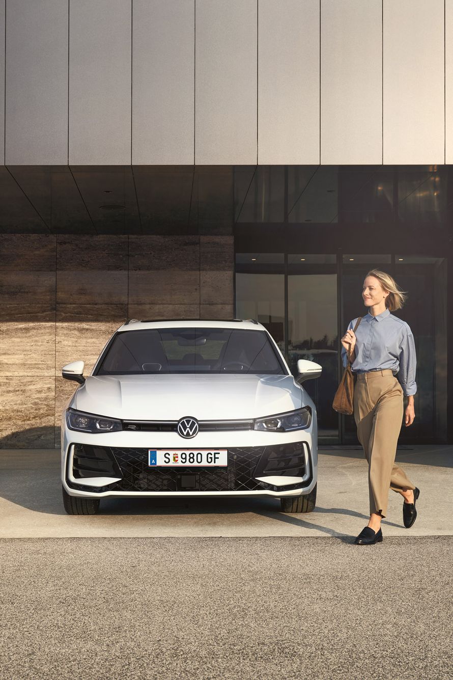 Frontansicht eines weißen VW Passat R-Line, eine Frau geht an ihm vorbei.