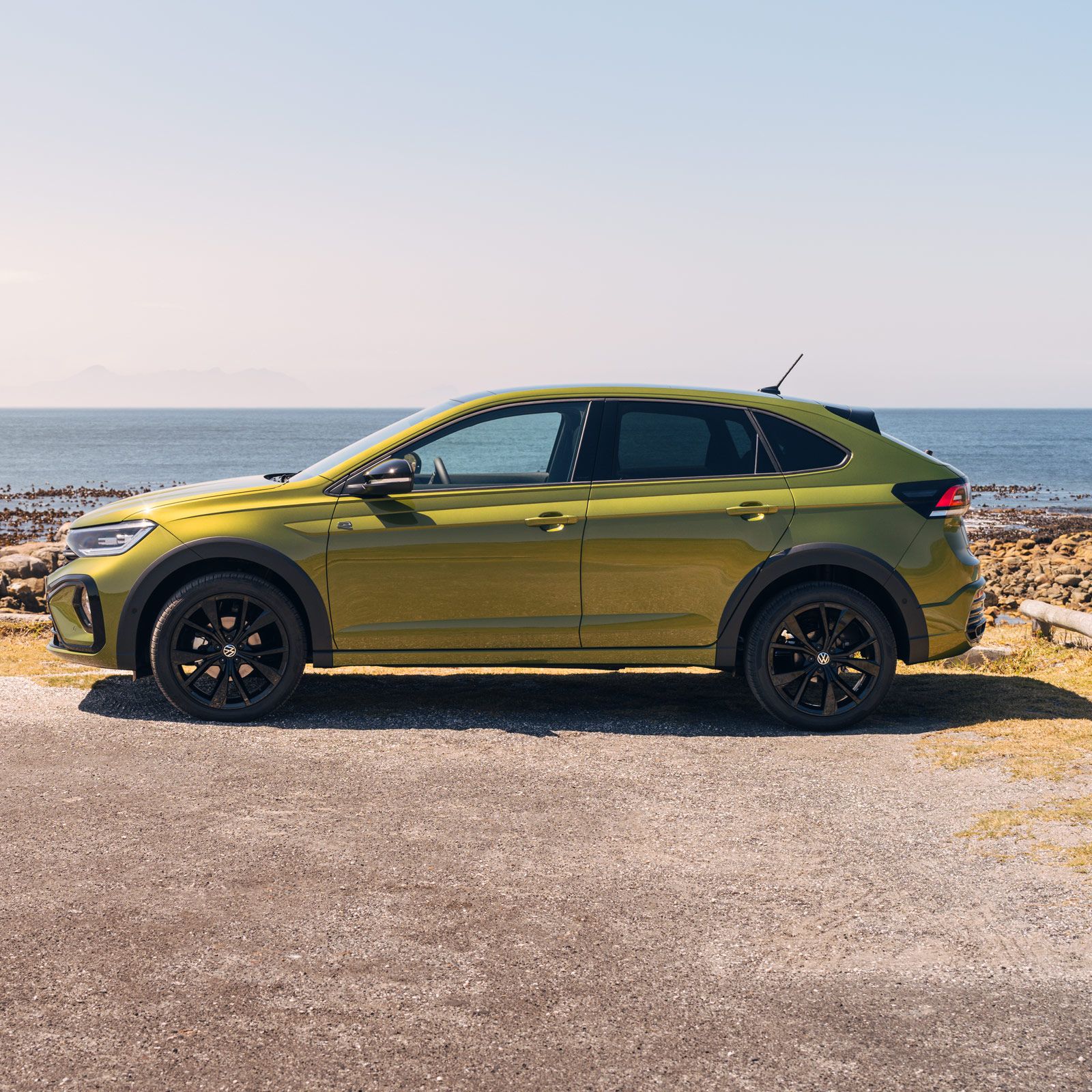 Ein grüner Taigo Black Edition steht seitlich am Meer mit sichtbaren Felsen am Ufer und einem klaren Himmel im Hintergrund. Das Auto hat schwarze Reifen und dunkle Fenster.