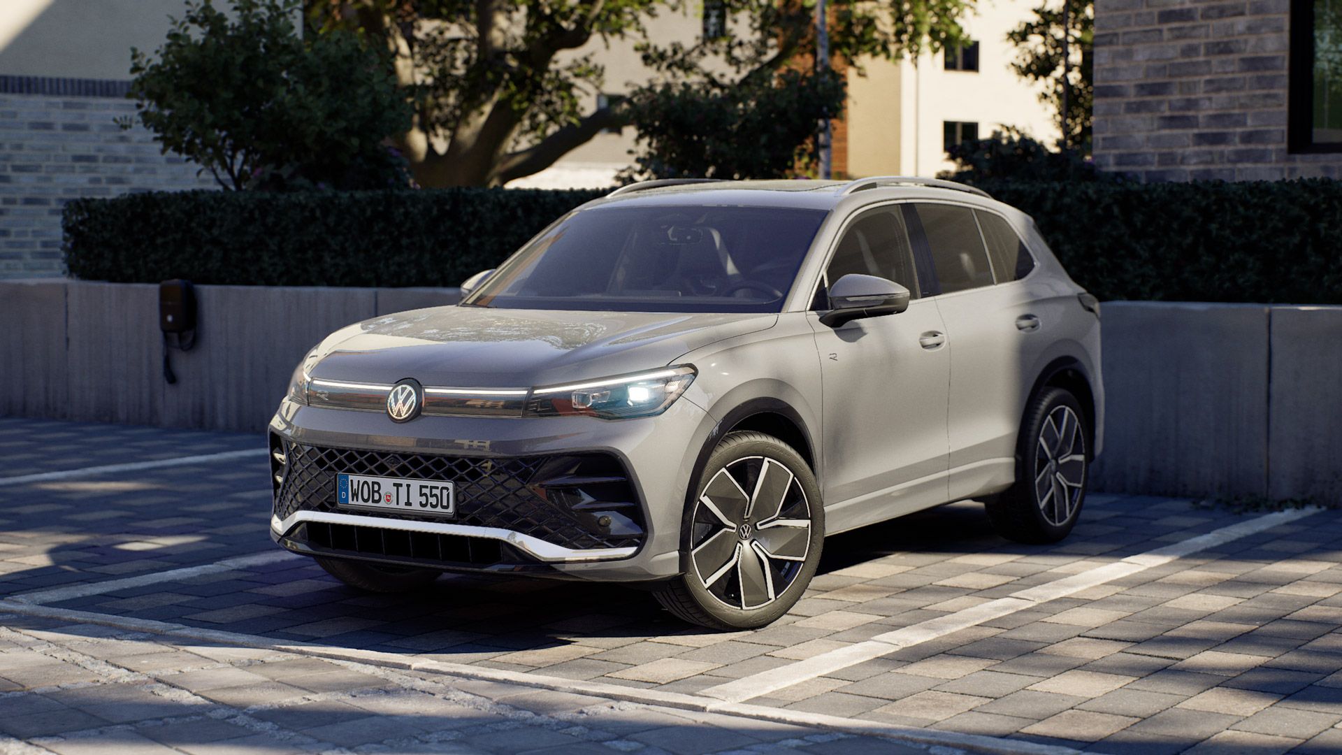 Silbergrauer VW Tiguan steht auf einem gepflasterten Parkplatz vor modernen Wohngebäuden. Das Auto ist schräg von vorn zu sehen, mit LED-Scheinwerfern und großen Alufelgen. Im Hintergrund sind Bäume und eine niedrige Betonmauer zu erkennen.