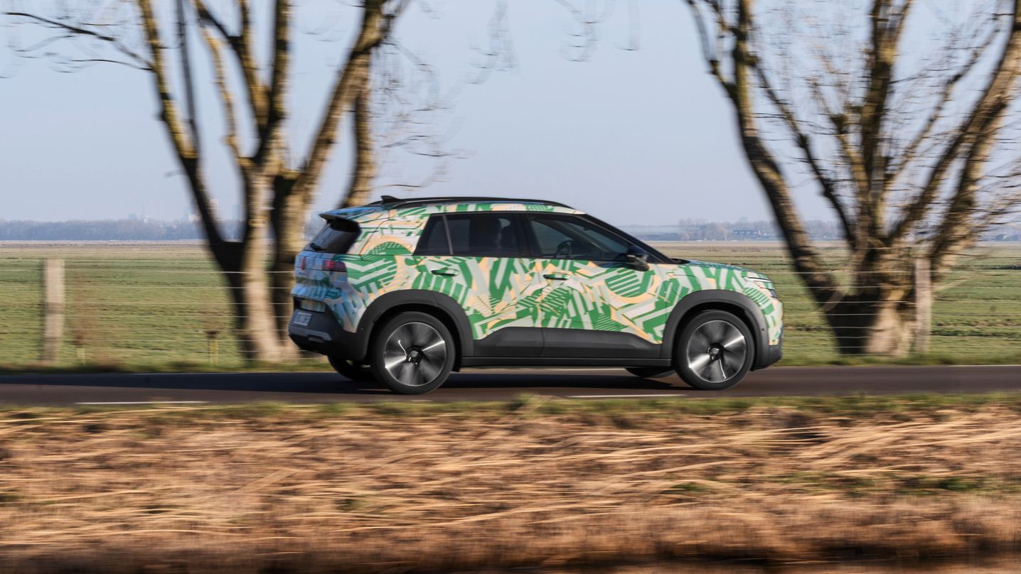 Ein SUV fährt auf einer ländlichen Straße vorbei an kahlen Bäumen und einem Wassergraben. Das Auto ist mit einem bunten, tropischen Muster bemalt. Im Hintergrund ist eine grüne Wiese und wolkenloser Himmel zu sehen.
