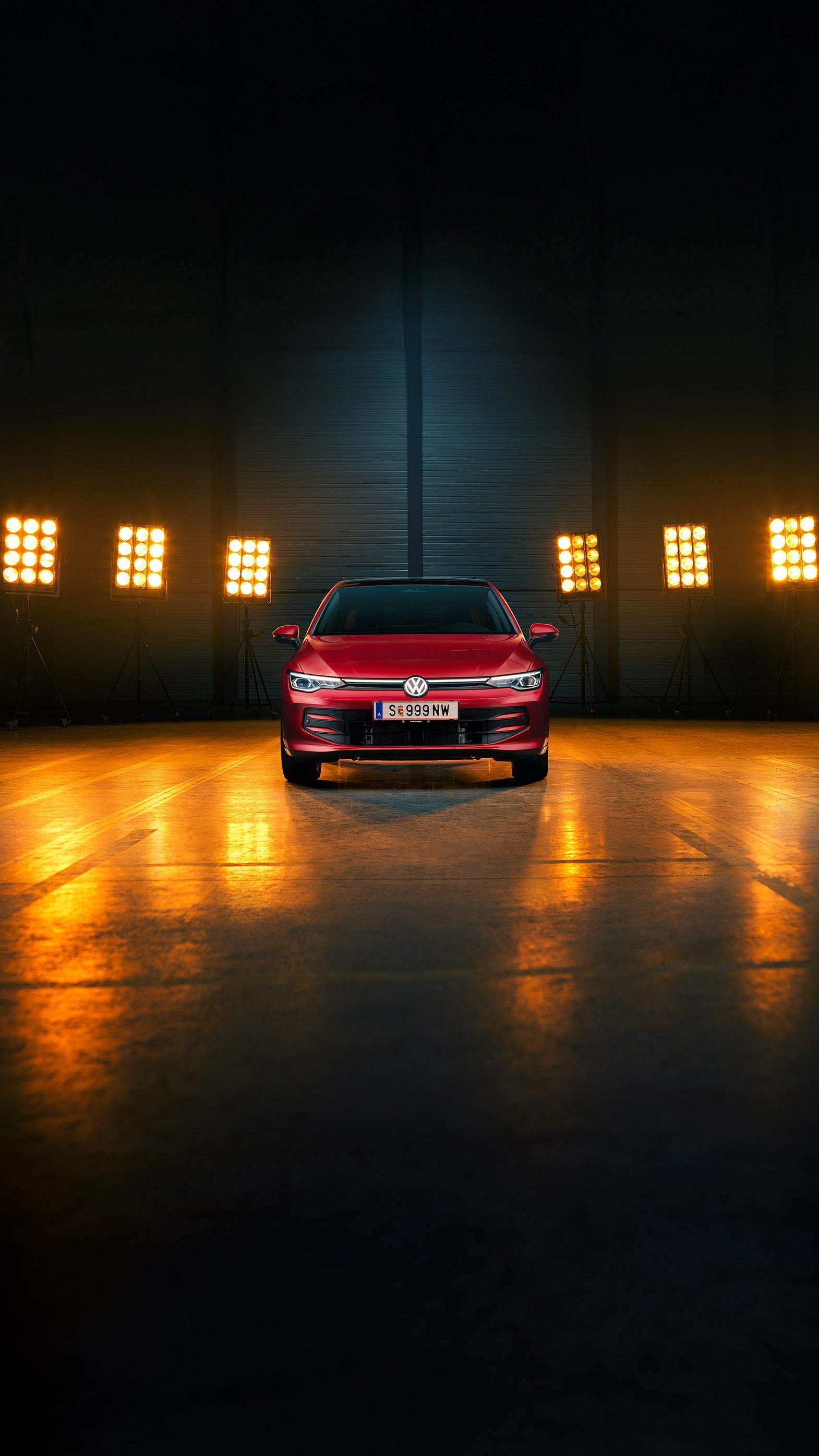 Ein roter VW Golf Falco Edition in Frontalansicht steht mittig im Bild in einer leeren Halle, Bühnenlichter strahlen das Fahrzeug von hinten an.