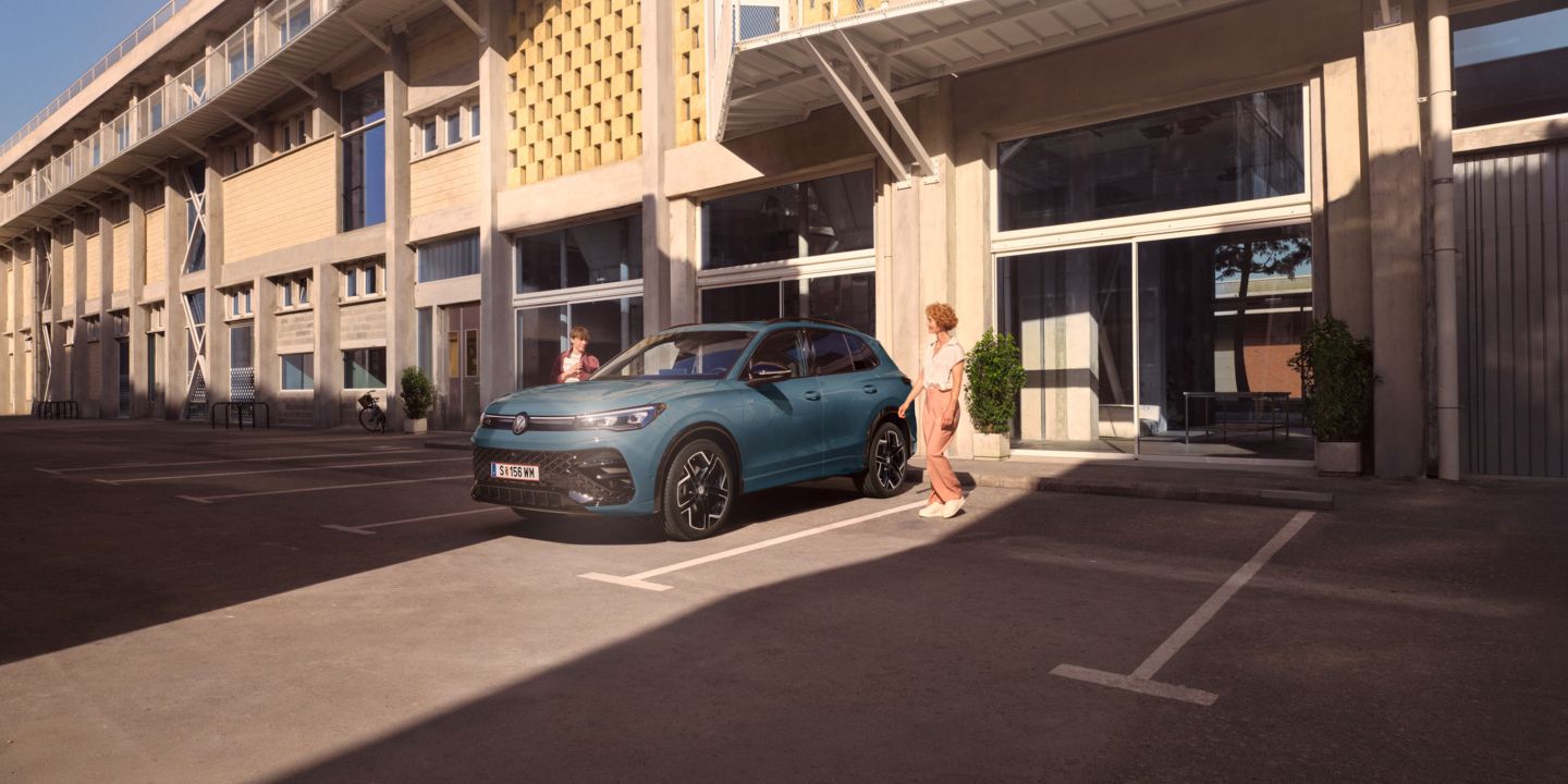 Eine junge Frau und ein junger Mann gehen zu ihrem roten Tiguan R-Line, der vor einem Bürogebäude auf einem Parkplatz steht.