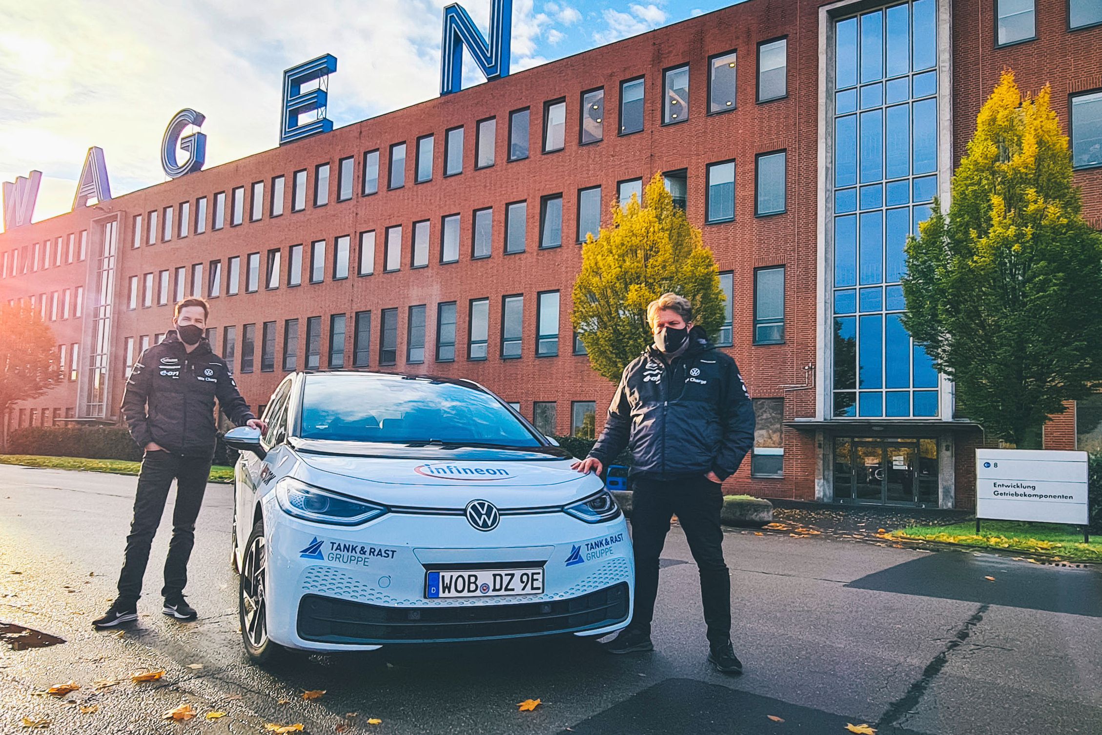 Zwei Personen stehen neben einem weißen ID.3 vor einem großen Backsteingebäude mit dem Schriftzug "VOLKSWAGEN" auf dem Dach. Die Sonne scheint, und das Wetter ist klar. Auf dem Boden liegen einige Herbstblätter.