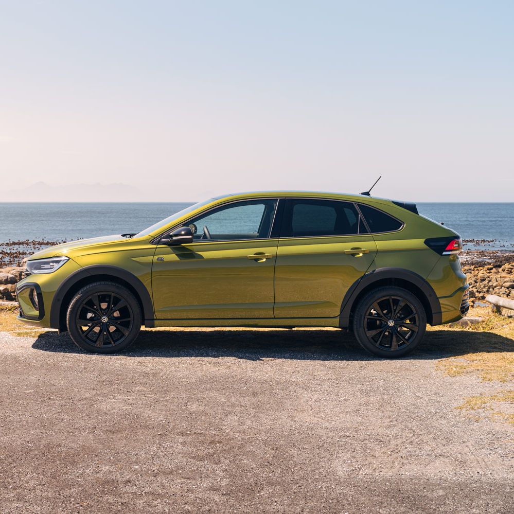 Ein grüner Taigo Black Edition steht seitlich am Meer mit sichtbaren Felsen am Ufer und einem klaren Himmel im Hintergrund. Das Auto hat schwarze Reifen und dunkle Fenster.