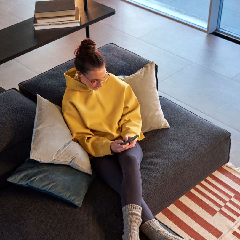 Person sitzt in einem modernen Wohnzimmer auf einem grauen Sofa und hält ein Handy in der Hand. Sie trägt einen gelben Hoodie, schwarze Leggings und graue Socken. Ein Bücherregal voller Bücher steht im Hintergrund.