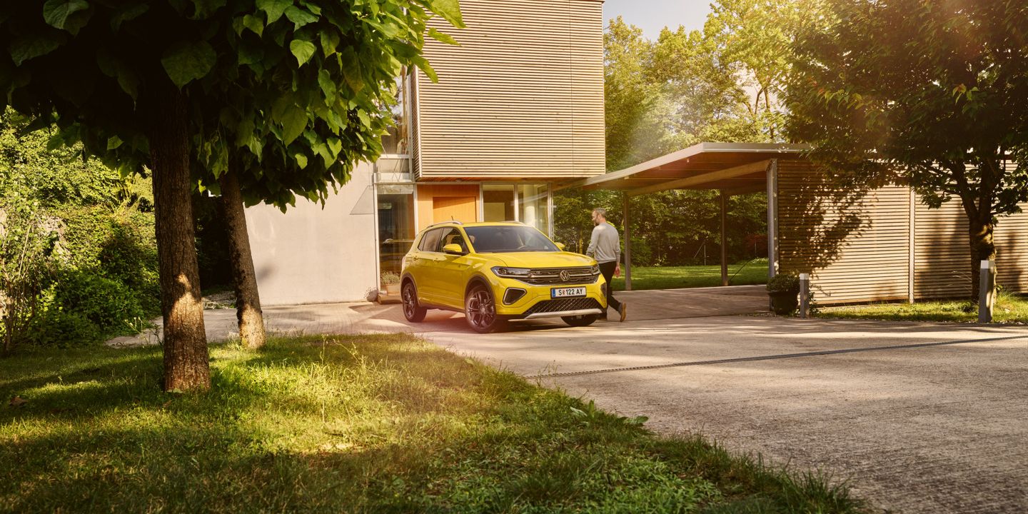Ein gelber VW T-Cross steht in der Auffahrt eines modernen Wohnhauses mit großen Bäumen.