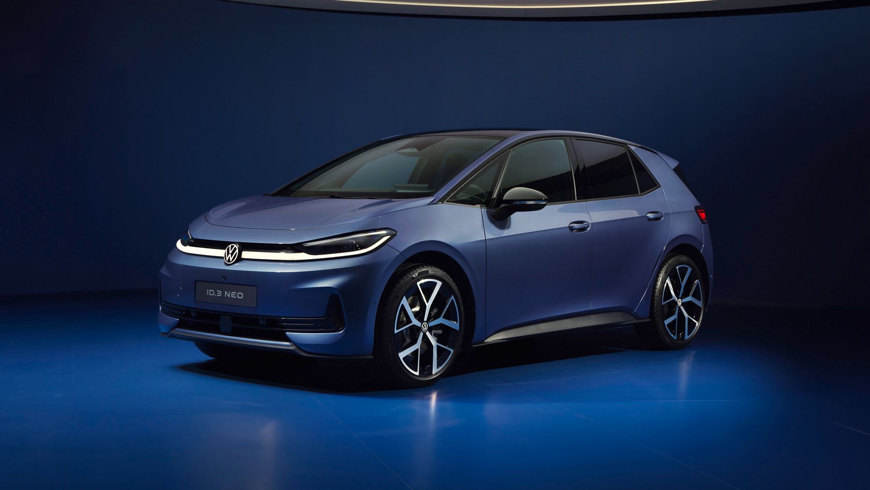 Ein dunkelblauer VW ID.3 NEO steht in einem futuristisch beleuchteten, komplett blauen Studio. Das Elektroauto ist schräg von vorne fotografiert, sodass Front und Fahrerseite gut sichtbar sind. Die Szene wirkt minimalistisch und hochwertig, mit weichem Licht und spiegelndem Boden.