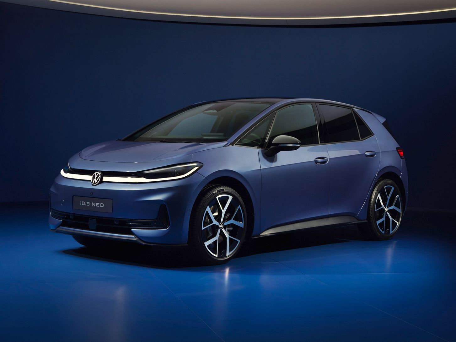 Ein dunkelblauer VW ID.3 NEO steht in einem futuristisch beleuchteten, komplett blauen Studio. Das Elektroauto ist schräg von vorne fotografiert, sodass Front und Fahrerseite gut sichtbar sind. Die Szene wirkt minimalistisch und hochwertig, mit weichem Licht und spiegelndem Boden.