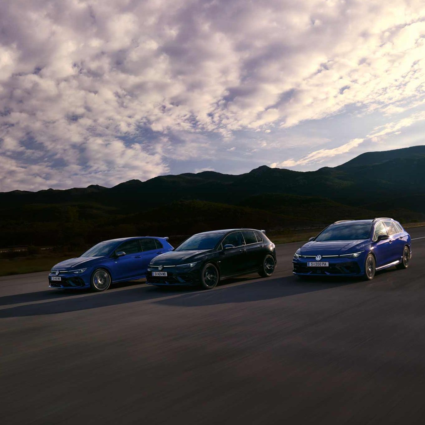 Drei VW Golf R fahren nebeneinander auf einer Straße bei Sonnenuntergang. Die Himmel zeigt einige Wolken und im Hintergrund sind Berge sichtbar. Die Farben der Autos sind blau, schwarz und dunkelblau.