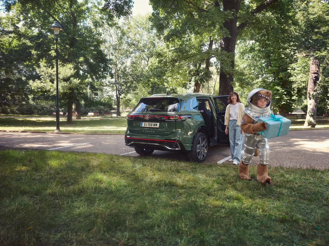 Eine Mutter steht neben einem grünen Volkswagen Tiguan und beobachtet ihr Kind, das mit einem Raumanzug bekleidet ist und ein Geschenk in den Händen hält.