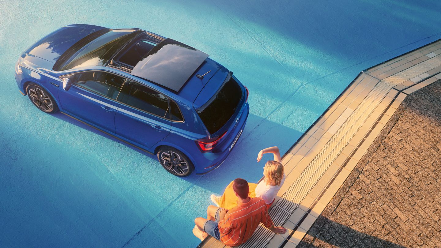  Blick von oben auf blauen VW Polo im Sonnenschein mit geöffnetem Panorama-Ausstell- /Schiebedach in einem leeren Pool. Zwei Personen sitzen am Rand des Pools.