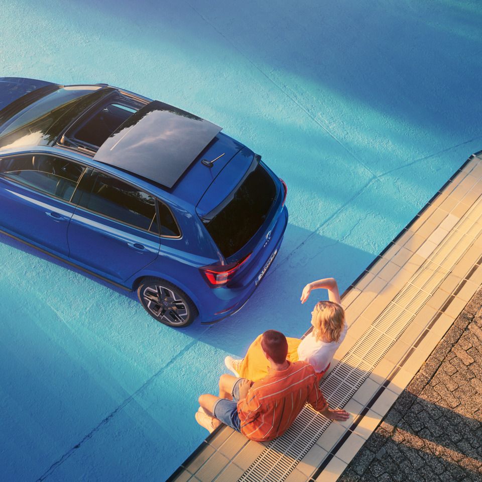 Blick von oben auf blauen VW Polo im Sonnenschein mit geöffnetem Panorama-Ausstell- /Schiebedach in einem leeren Pool. Zwei Personen sitzen am Rand des Pools.
