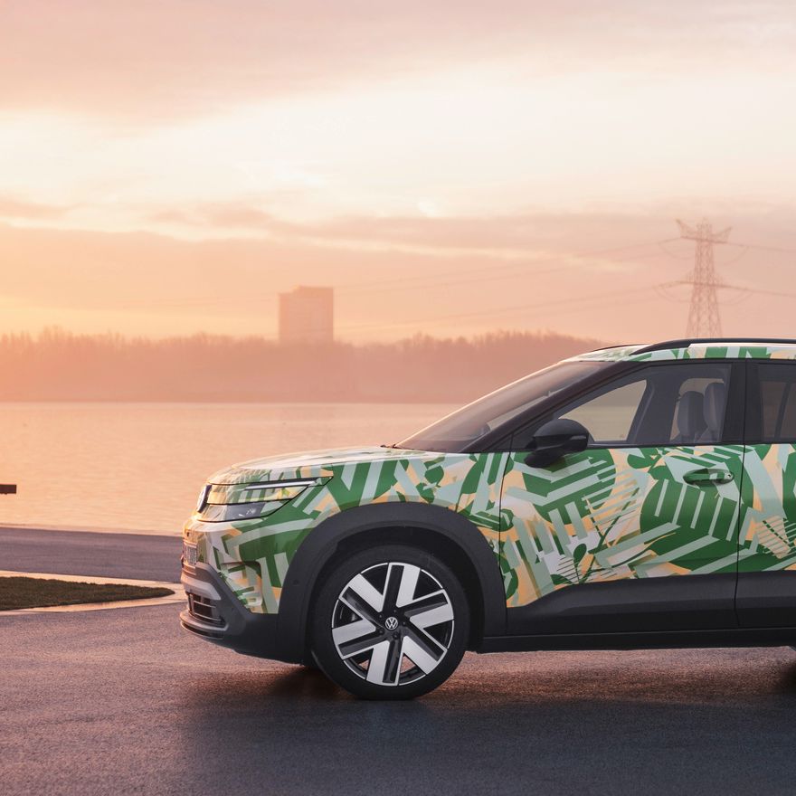 Ein SUV mit einer grünen und weißen Tarnlackierung steht auf einem asphaltierten Platz vor einem Gewässer bei Sonnenuntergang. Im Hintergrund sind Strommasten und Gebäude zu sehen. Der Himmel ist leicht bewölkt und in warmen Farben gehalten.