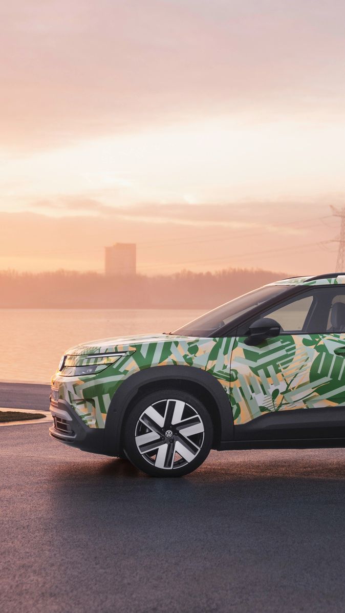 Ein SUV mit einer grünen und weißen Tarnlackierung steht auf einem asphaltierten Platz vor einem Gewässer bei Sonnenuntergang. Im Hintergrund sind Strommasten und Gebäude zu sehen. Der Himmel ist leicht bewölkt und in warmen Farben gehalten.