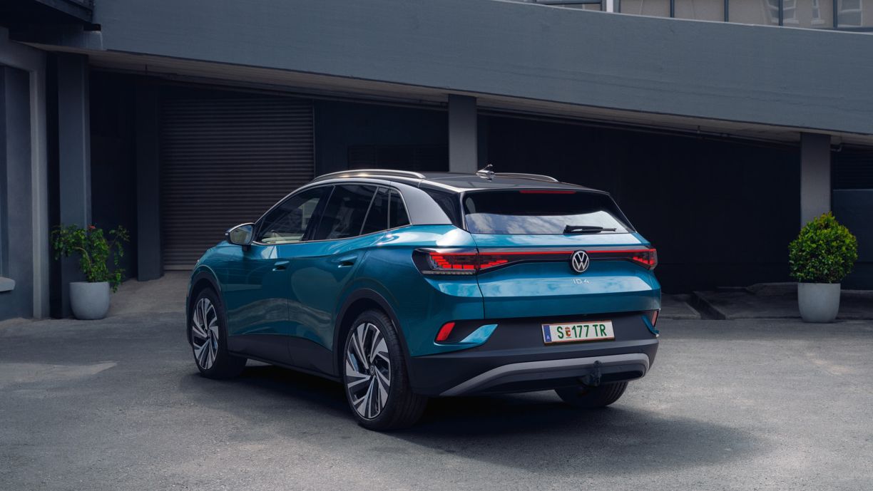 Ein blauer Volkswagen ID.4 steht geparkt in einem modernen, städtischen Umfeld. Das Fahrzeug zeigt seine Rückansicht. Im Hintergrund sind Gebäude aus Glas und Beton zu sehen.
