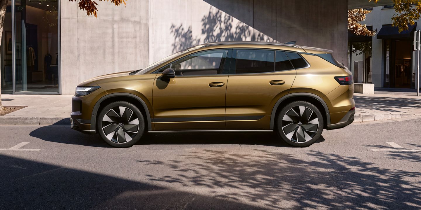 Seitenansicht eines oliv-bronzefarbenen VW ID.4 Reskin geparkt vor einem modernen Wohngebäude.
