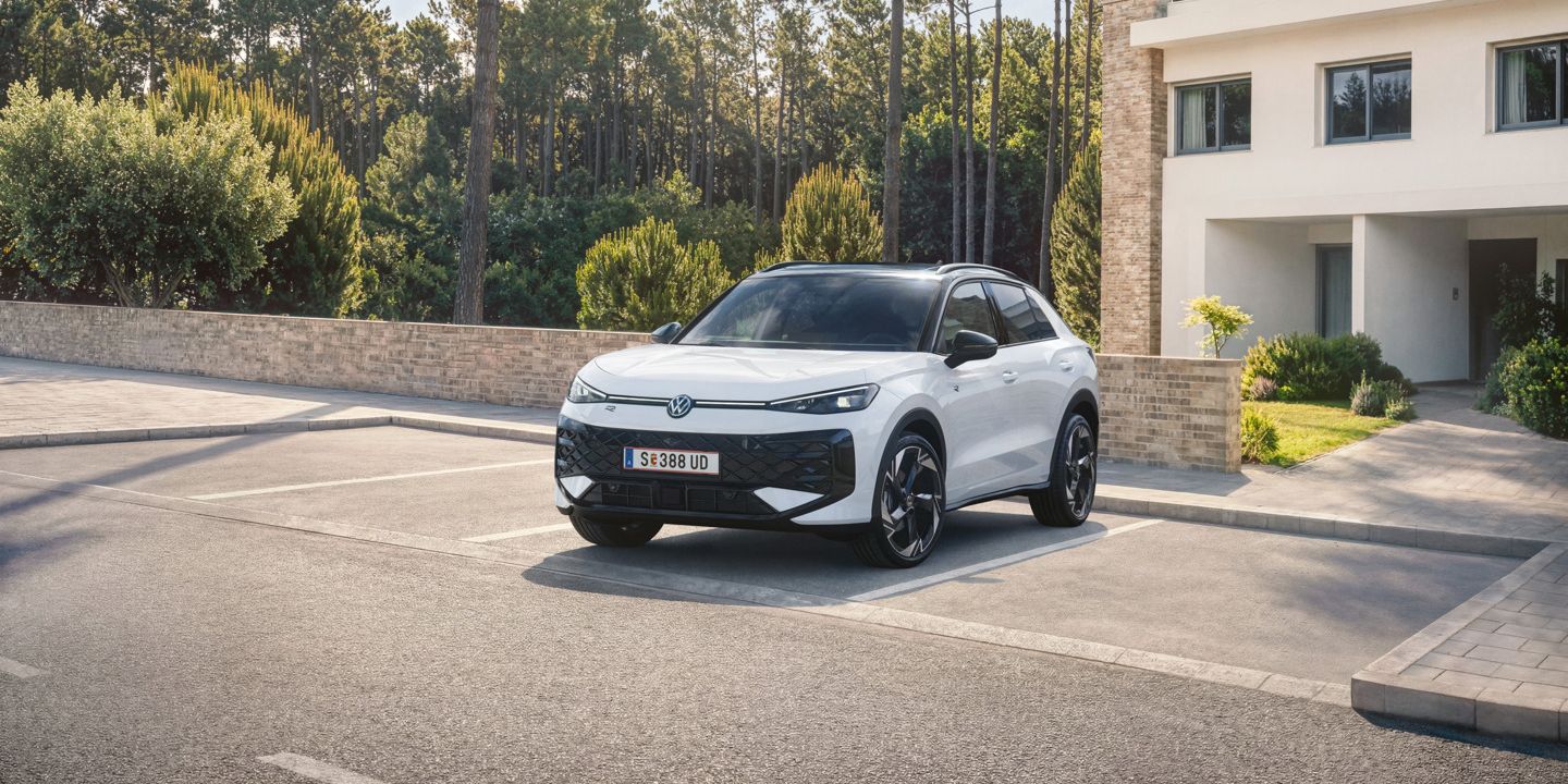Ein weißer VW T-Roc Hybrid steht auf einem Parkplatz vor einem Wohnhaus am Wald, Blick auf die Front.