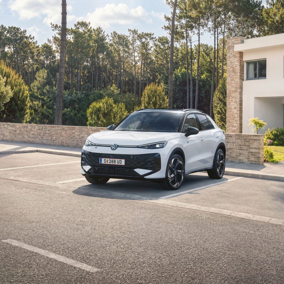 Ein weißer VW T-Roc Hybrid steht auf einem Parkplatz vor einem Wohnhaus am Wald, Blick auf die Front.