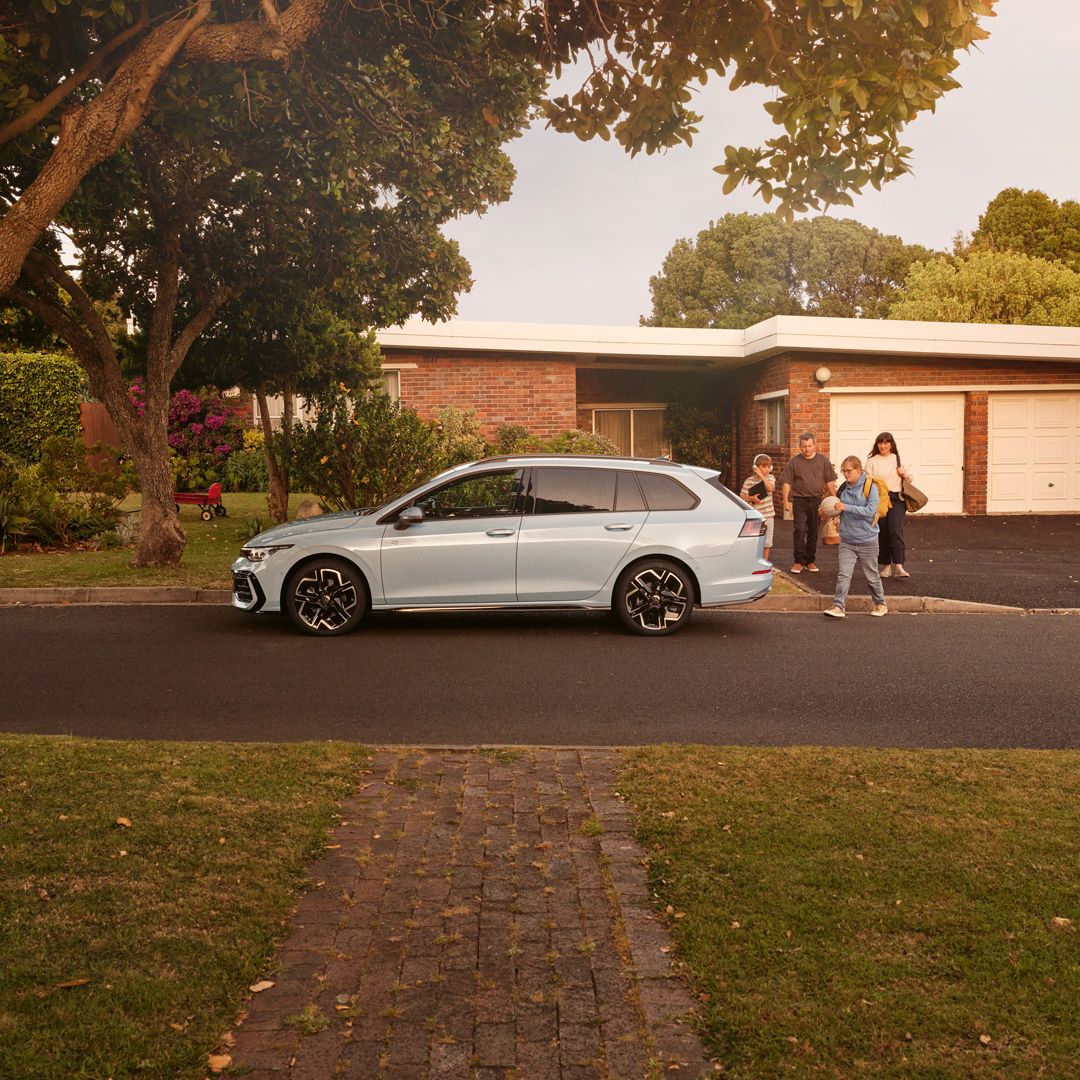 Ein weißer VW Golf Variant parkt vor einem Bungalow in einer Wohnstraße mit Bäumen. 