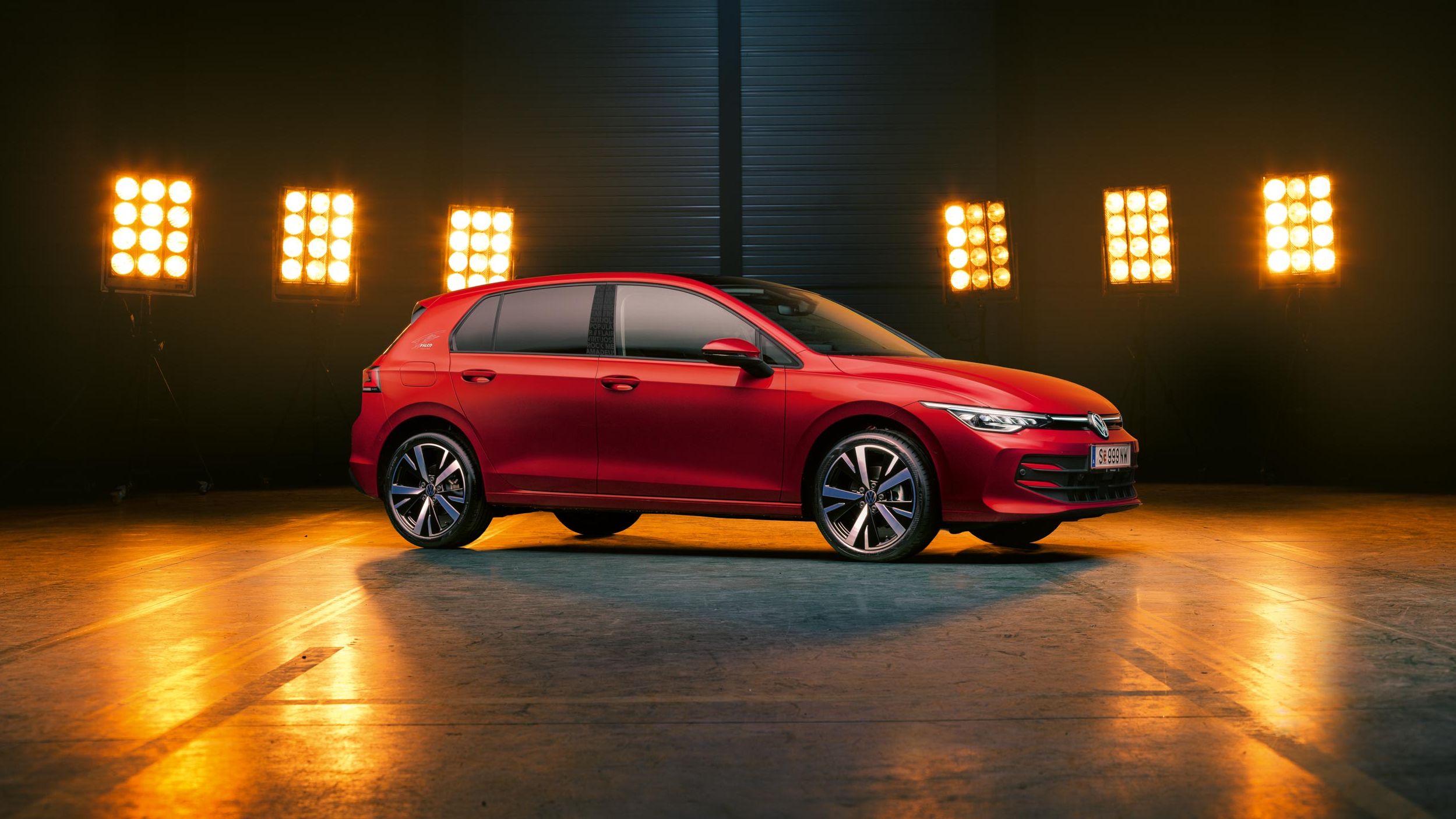 Seitenansicht auf einen roten VW Golf Falco Edition, der in einer leeren Halle steht, dahinter wird er von Scheinwerfern beleuchtet.