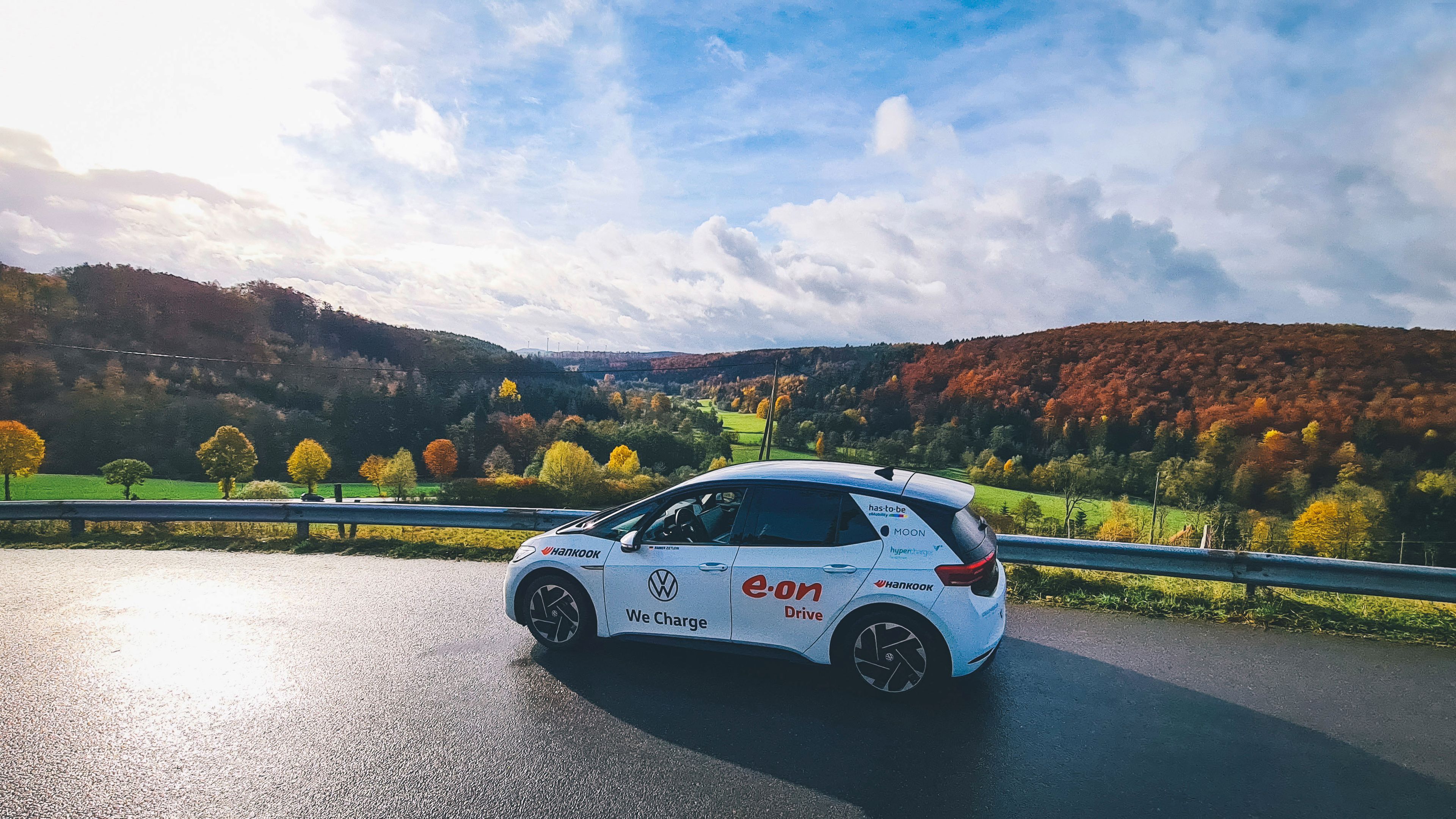 Ein ID.3 von Volkswagen fährt an einer malerischen Landschaft mit grünen Feldern und bunt gefärbten Bäumen vorbei. Der Himmel ist leicht bewölkt und die Sonne scheint durch. Auf dem Auto steht "e-on Drive" und "We Charge".