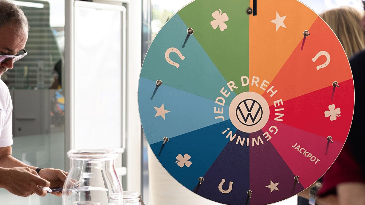 Eine Person dreht ein großes, buntes Glücksrad, das verschiedene Symbole und den Schriftzug "GEWINN! JEDER DREH EIN JACKPOT" zeigt. Daneben steht ein Tisch mit Gläsern, in denen kleine Gegenstände liegen. Eine andere Person schaut zu und hält ein Getränk.
