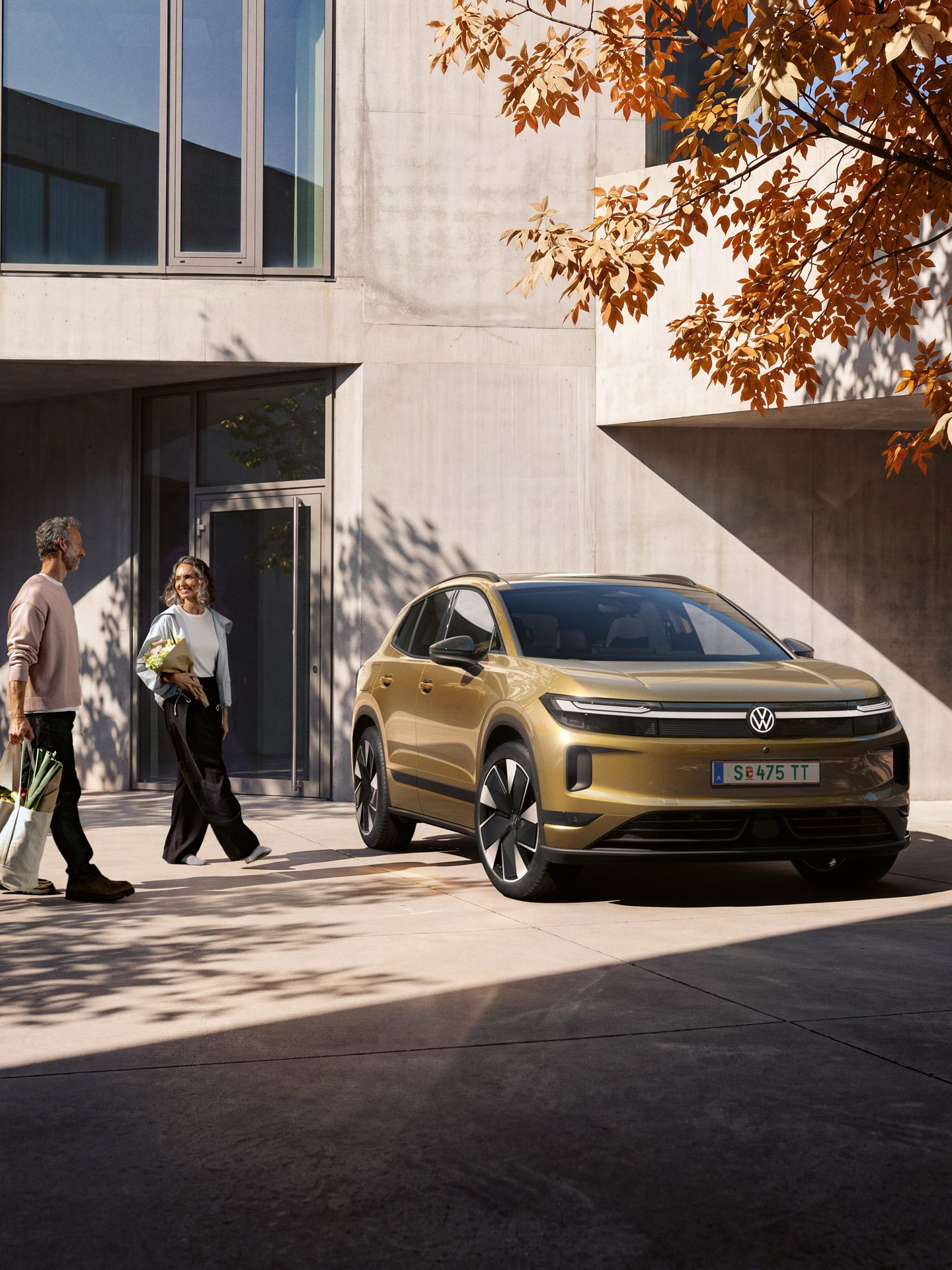 Ein Mann und eine Frau gehen mit Einkäufen beladen auf einen oliv-bronzefarbenen ID.4 Reskin zu, der vor einem modernen Gebäude parkt.