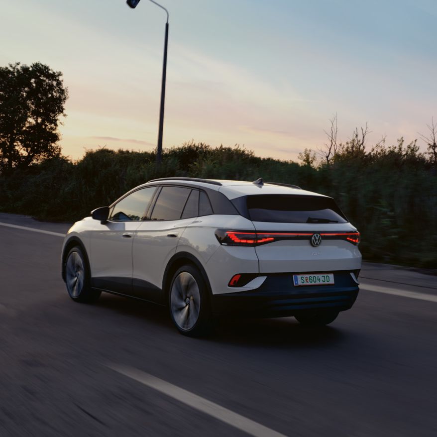Weißer VW ID.4 in Heckansicht fährt auf einer Landstraße bei Sonnenuntergang.