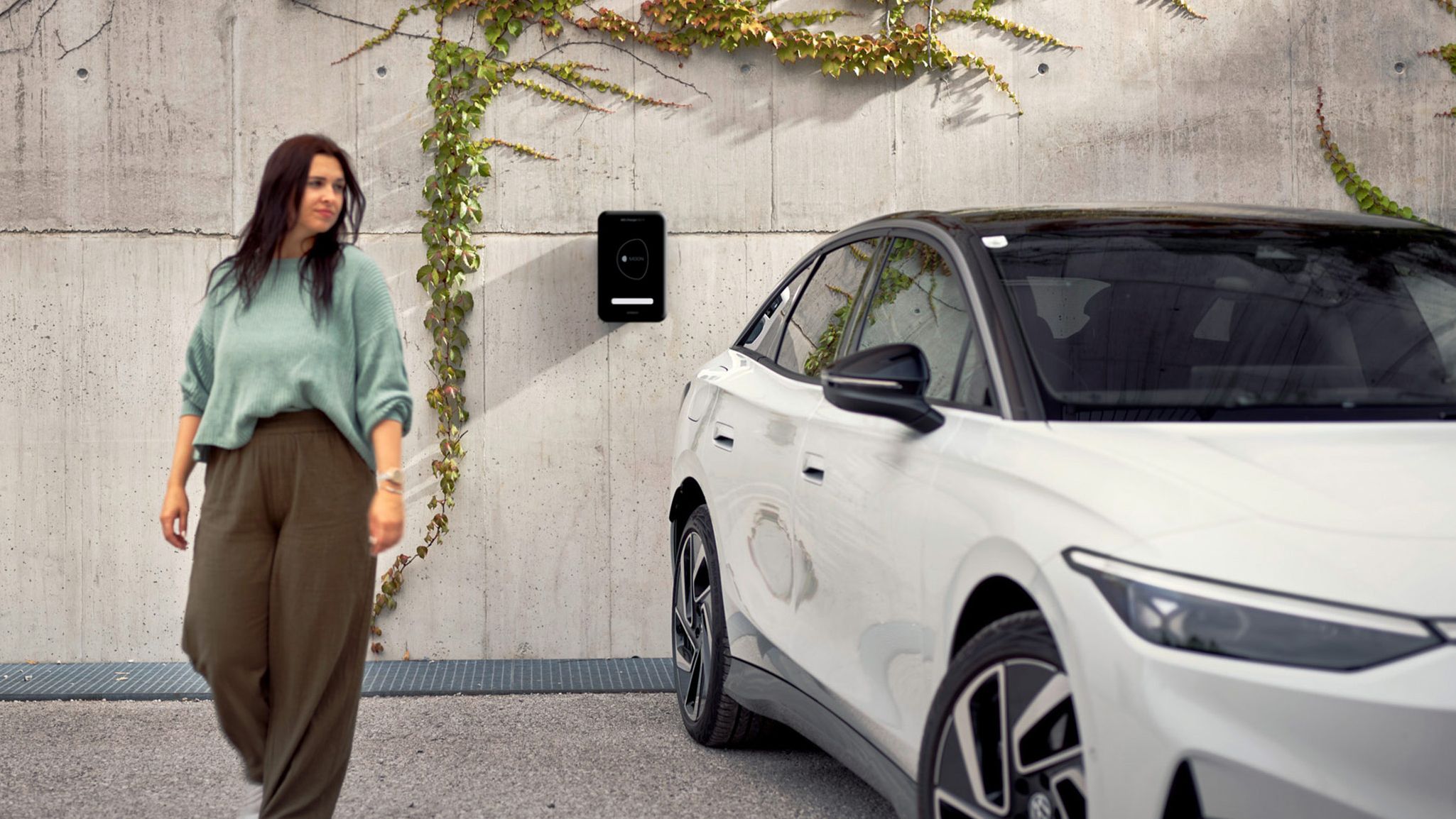 Eine Frau geht in der Nähe eines weißen ID.7, das an einer Wand mit einer Ladestation und Kletterpflanzen geparkt ist. Die Umgebung wirkt urban und modern.