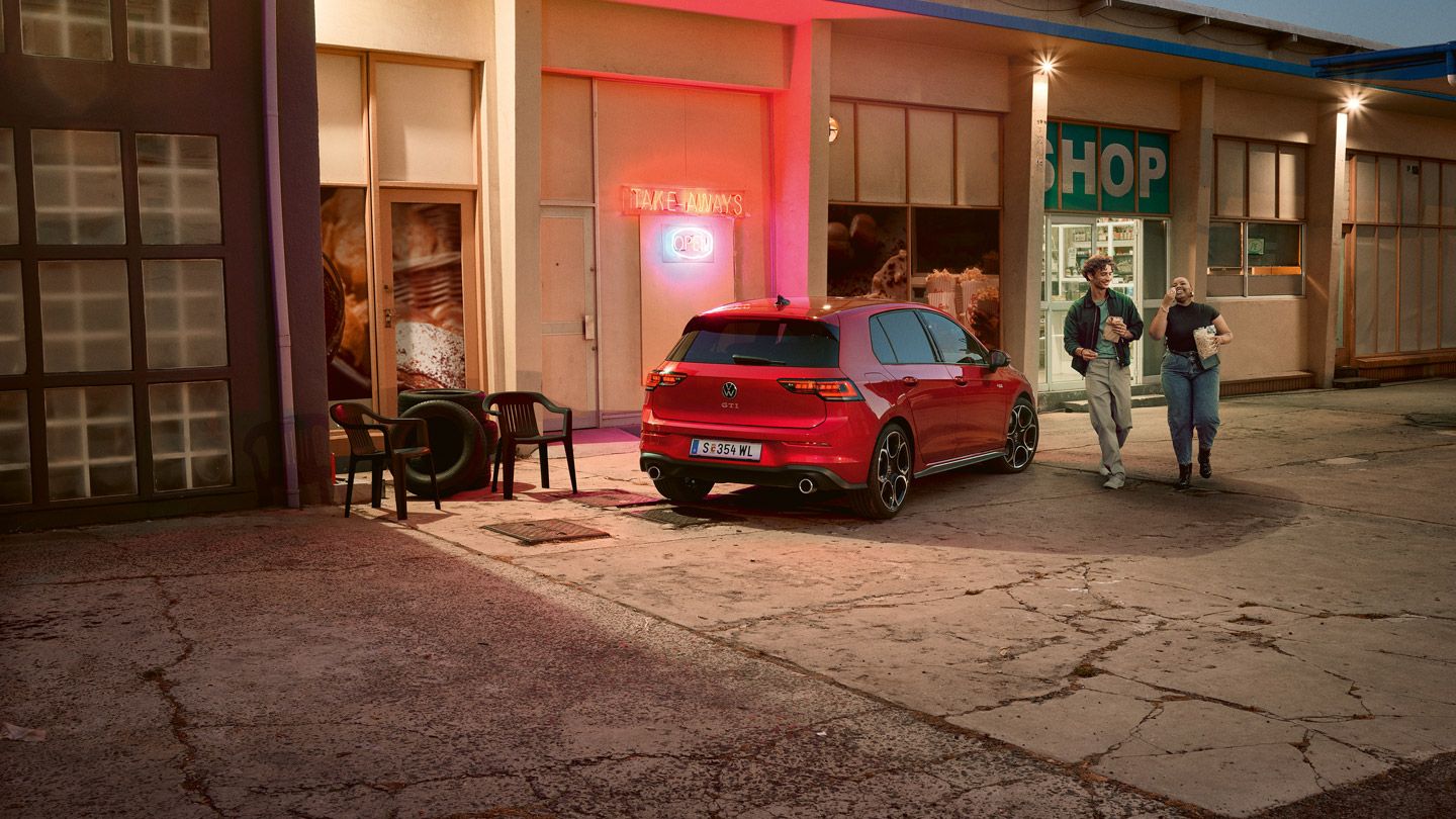 Roter VW Golf GTI steht abends vor einem Supermarkt, zwei junge Leute gehen mit Einkäufen daran vorbei.